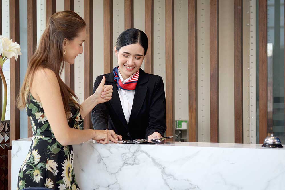 Beautiful smiling Asian receptionist suggests travel program for tourist woman customer, female staff at reception counter desk provides information, guest booking tour at a hotel on holiday vacation